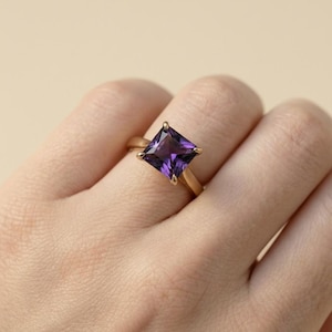 May include: A gold ring featuring a square-cut, deep purple gemstone. The ring's band is a warm gold color, and the gemstone is set in a four-prong setting. The ring is shown on a person's finger.