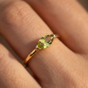 May include: A gold ring with a marquise-cut, light green gemstone. The gemstone is set in a gold band with a delicate, minimalist design. The ring is shown on a finger.