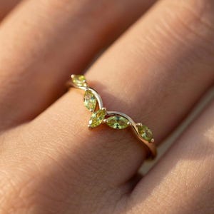 May include: A gold-colored ring with a curved design, featuring several oval-shaped, light green gemstones. The ring is worn on a finger, showcasing the delicate details of the jewelry.