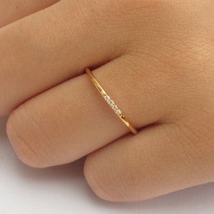 Può includere: Un delicato anello d'oro con una fascia sottile, ornato da una fila di piccole pietre preziose scintillanti. L'anello è mostrato su un dito, evidenziando il suo design elegante e minimalista. Perfetto per l'uso quotidiano o occasioni speciali.