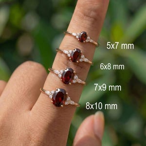 May include: Four gold rings with oval-cut dark red garnet center stones and small white accent stones on the bands. The rings are labeled with sizes 5x7 mm, 6x8 mm, 7x9 mm, and 8x10 mm, showing the varying dimensions of the garnet gemstones.