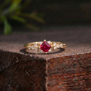 May include: A gold ring featuring a central princess-cut red gemstone flanked by three small round white stones on each side. The band is thin and polished, showcasing a symmetrical arrangement of the vibrant red stone and clear accent stones.