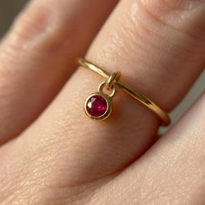 以下が含まれることがあります： 小さな丸いルビーのペンダントが付いた繊細なゴールドリング。リングは細く磨かれたバンドで、ルビーは金のベゼルにセットされています。鮮やかな赤い宝石は、温かみのあるゴールドの色と対照的で、シンプルでありながらエレガントなジュエリーです。