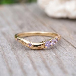 May include: A gold curved band ring featuring three round purple gemstones set on one side, with additional small purple stones and engraved star motifs scattered along the polished metal surface of the band.
