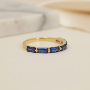 May include: A gold band ring featuring a series of rectangular, deep blue gemstones. The ring is set against a soft, neutral background, highlighting the contrast between the gold and the blue stones. The ring's design is simple and elegant.