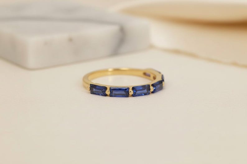 May include: A gold band ring featuring a series of rectangular, deep blue gemstones. The ring is set against a soft, neutral background, highlighting the contrast between the gold and the blue stones. The ring's design is simple and elegant.