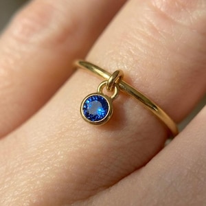 Könnte beinhalten: Ein goldener Ring mit einem kleinen, runden, tiefblauen Edelsteinanhänger. Der Ring ist schmal und schlicht, wobei der Edelstein an einer kleinen Schlaufe hängt. Der Edelstein hat eine leuchtend blaue Farbe.