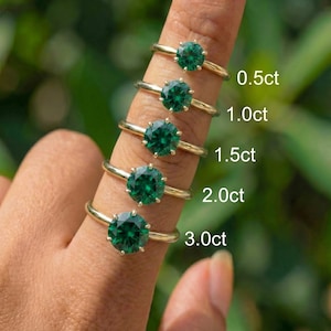 Puede incluir: Una colección de anillos dorados con piedras preciosas verdes con corte esmeralda, exhibidos en un dedo. Los anillos están etiquetados con pesos en quilates: 0,5 ct, 1,0 ct, 1,5 ct, 2,0 ct y 3,0 ct. El fondo es verde suave.
