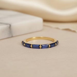 May include: A gold band ring with rectangular, deep blue gemstones set around the band. The ring is displayed on a neutral-toned surface with a soft, blurred background. The ring is a piece of jewelry.