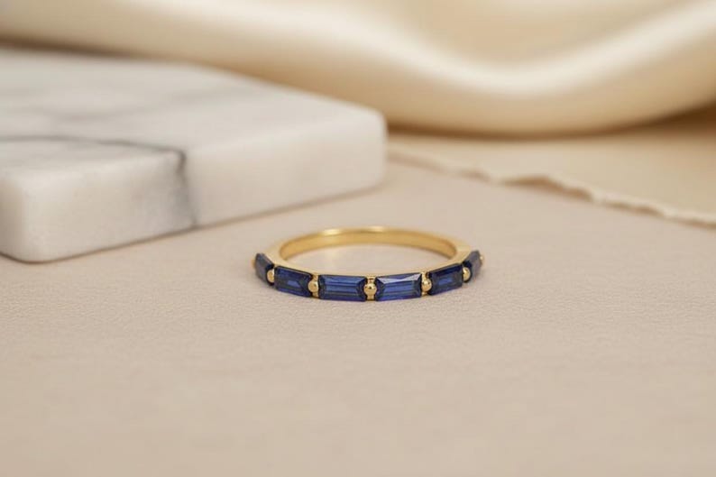May include: A gold band ring with rectangular, deep blue gemstones set around the band. The ring is displayed on a neutral-toned surface with a soft, blurred background. The ring is a piece of jewelry.