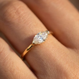 May include: A close-up of a gold ring featuring a marquise-cut diamond. The diamond is set in a gold band, and the ring is on a finger. The diamond sparkles in the light.