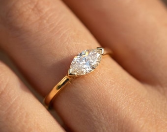 May include: A close-up of a gold ring featuring a marquise-cut diamond. The diamond is set in a gold band, and the ring is on a finger. The diamond sparkles in the light.