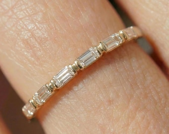 May include: A delicate gold ring featuring a series of rectangular, clear gemstones. The ring's band is a warm gold color, and the gemstones are set in a linear pattern. The ring is designed to be worn on a finger.
