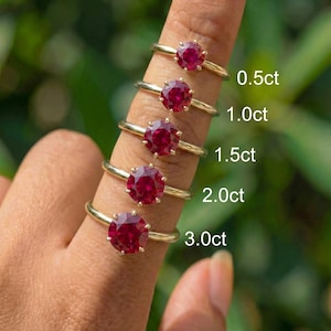 May include: Five gold rings with round, deep red gemstones are displayed on a finger. The rings are labeled with their carat weights: 0.5ct, 1.0ct, 1.5ct, 2.0ct, and 3.0ct. The background is a soft green.