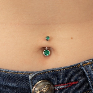 May include: A belly button piercing with two green gem stones. The piercing is on a person wearing blue jeans.