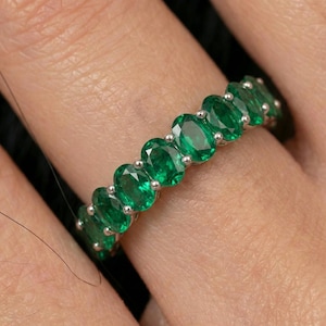 Puede incluir: Un anillo de plata con una fila de esmeraldas de corte ovalado. Las vibrantes piedras preciosas verdes están engastadas juntas, creando una banda continua de color. El anillo se muestra en un dedo, resaltando el diseño y el brillo de la joya.