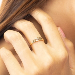May include: A gold-colored ring with a mountain-peak design. The ring is worn on a finger, with the hand and arm visible. The ring's design features a series of triangular shapes, creating a mountain silhouette. The ring is approximately 0.75 inches in diameter.