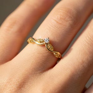 May include: A gold-colored ring with a curved design. The ring features a central, clear gemstone and smaller yellow gemstones along the band. The ring is on a finger, with the focus on the jewelry.