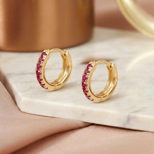 May include: A pair of small gold hoop earrings featuring a row of vibrant, round-cut pinkish-red gemstones set along the front-facing outer edge of each hoop. The earrings have a polished metallic finish and a secure hinged closure mechanism.