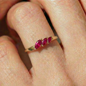 May include: A gold ring featuring three marquise-cut, vibrant red gemstones. The gemstones are set in a row, creating a delicate and elegant design. The ring's band is a simple, polished gold.