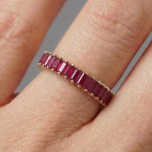 May include: A gold ring featuring a row of rectangular, deep red gemstones. The gemstones are set closely together, creating a continuous band of color. The ring is shown on a finger.