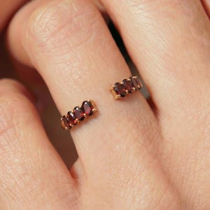 May include: A gold-toned open ring featuring a row of oval-cut, deep red gemstones. The ring is split, with a section of gemstones on each side of the finger. The gemstones are set in a gold band.