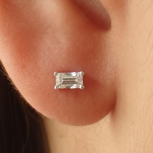 May include: A close-up of a small, rectangular diamond stud earring. The diamond is clear and has a baguette cut. The earring is set in a silver or white metal. The earring is on a person's ear.