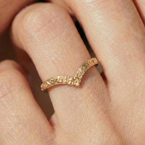 Puede incluir: Un anillo dorado con un diseño floral. El anillo tiene forma de V y está adornado con intrincados patrones de flores y hojas. El anillo se lleva en un dedo.