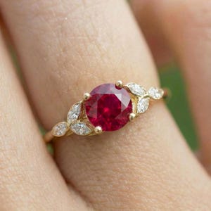 May include: A gold ring featuring a large, round, deep red gemstone as the centerpiece. The band is adorned with smaller, clear gemstones in a leaf-like pattern on either side of the central stone. The ring is set against a blurred background.