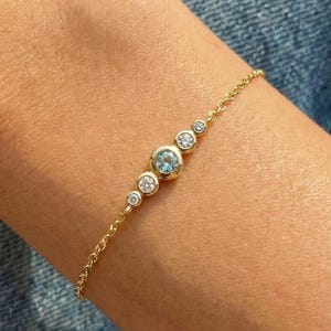 Może przedstawiać: Delikatna złota bransoletka z serią okrągłych kamieni szlachetnych. Bransoletka ma centralny jasnoniebieski kamień szlachetny, otoczony mniejszymi, przezroczystymi kamieniami szlachetnymi. Bransoletka ma około 18 cm długości.