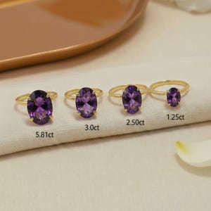 Puede incluir: Cuatro anillos de oro con piedras preciosas de amatista púrpura ovaladas de diferentes tamaños. Los anillos están etiquetados con sus pesos en quilates: 5,81 ct, 3,0 ct, 2,50 ct y 1,25 ct. Los anillos se muestran sobre una tela blanca.