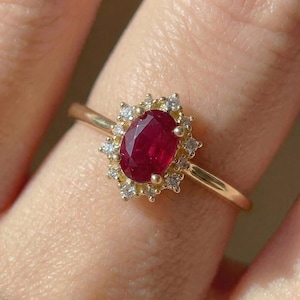 Op de afbeelding: Een gouden ring met een ovale robijn omringd door kleine diamanten. De dieprode robijn wordt geaccentueerd door een kroonvormige zetting. De ring heeft een smalle, gouden band.