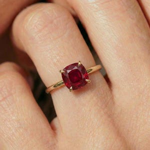 May include: A gold ring featuring a large, square-cut, deep red gemstone. The gemstone is held in place by four prongs. The ring's band is a thin, polished gold. The ring is shown on a finger.