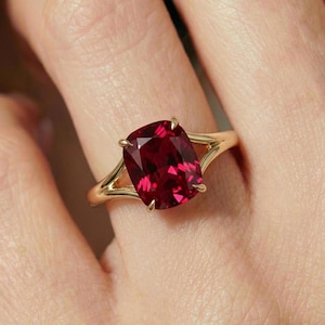 Pode incluir: Um anel de ouro com uma grande pedra preciosa vermelha escura com corte almofada. A banda do anel tem um design dividido, com a pedra preciosa presa por quatro garras. O anel é mostrado num dedo.