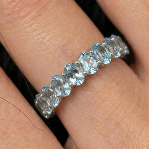 May include: A silver ring set with a row of oval-shaped, pale blue gemstones. The gems are closely set, forming a continuous band. The ring is displayed on a finger, showcasing the design and colour of the jewellery.