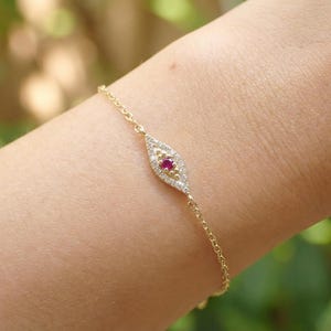 May include: A delicate gold bracelet featuring a central, eye-shaped charm. The charm is adorned with a vibrant red gemstone at its center, surrounded by smaller, sparkling white stones. The bracelet's chain is a fine, linked design.