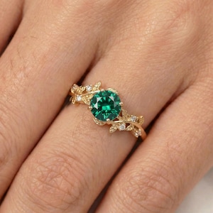 May include: A gold ring featuring a large, round, emerald-green gemstone. The ring's band is adorned with delicate leaf and small diamond accents. The ring is a nature-inspired design, perfect for a special occasion.