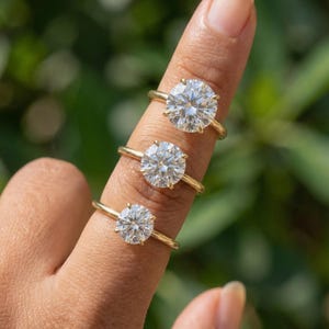 May include: Three gold rings with large, round, clear gemstones are displayed on a finger. The rings have a simple, classic design with a thin gold band. The gemstones sparkle brightly against a blurred green background.