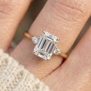 May include: A gold engagement ring featuring a large emerald-cut diamond as the centerpiece, flanked by two smaller round diamonds. The ring's band is a delicate gold, and the diamonds sparkle brightly.