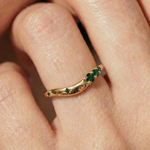 May include: A gold curved ring featuring three round green gemstones set in a row, accented by small engraved star motifs along the polished metal band. The band follows a gentle arc shape, showcasing a smooth, reflective finish throughout.