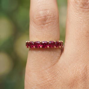 May include: A gold ring featuring a row of seven square-cut, vibrant red gemstones. The ring is set in a yellow gold band, creating a classic and elegant design. The gemstones are closely set, showcasing a continuous line of color.