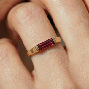 May include: A gold ring featuring a rectangular, deep red gemstone. The ring's band is a simple, polished gold, with the gemstone set flush within the design. The gemstone's color is a rich, vibrant red.