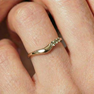 May include: A gold ring with a curved, wave-like band featuring three small, light green round-cut gemstones set in a row along the upper curve of the metal shank, which has a polished, smooth, and reflective surface finish.