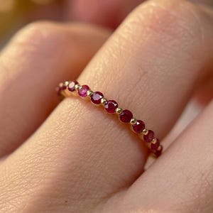 May include: A gold ring featuring a series of small, vibrant red gemstones. The gemstones are evenly spaced around the band, creating a continuous line of color. The ring is shown on a finger, highlighting its delicate design.