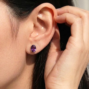 May include: Oval-shaped amethyst stud earring with a gold setting. The purple gemstone is set in a gold bezel. The earring is worn on the ear of a person.