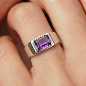 May include: A silver ring with a rectangular, deep purple gemstone. The ring has a simple, modern design with a polished finish. The gemstone is set in a bezel setting, and the band is wide and flat.