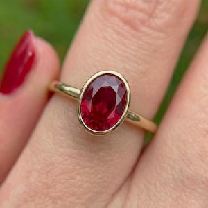 May include: A gold ring featuring a large, oval-cut ruby gemstone. The ruby is a deep red color and is set in a bezel setting. The ring's band is a simple, polished gold.