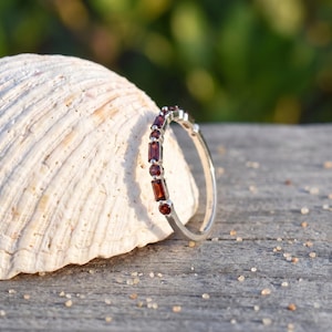 Op de afbeelding: Een zilveren ring met een reeks afwisselend rechthoekige en ronde dieprode granaat edelstenen langs de band. De ring staat rechtop tegen een getextureerde, gebroken witte schelp, waardoor de metalen afwerking en stenen zichtbaar zijn.