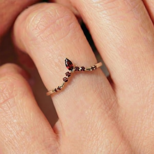 May include: A gold-colored ring with a marquise-cut red gemstone and several smaller round red gemstones. The ring is curved and sits on a finger.