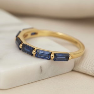 May include: A gold ring with a row of rectangular, deep blue gemstones. The ring is set on a white marble surface, with a soft, neutral-colored fabric in the background. The ring's band is a polished gold.